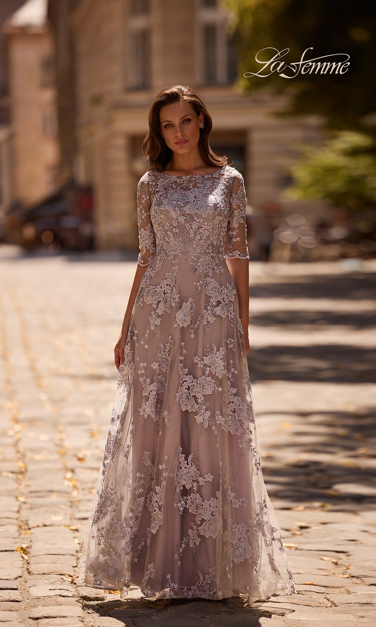 Woman stands outdoors in the Long Formal Dress: La Femme 27854, a floor-length lavender/gray gown with floral lace embroidery and three-quarter sleeves. Cobblestone street behind her.